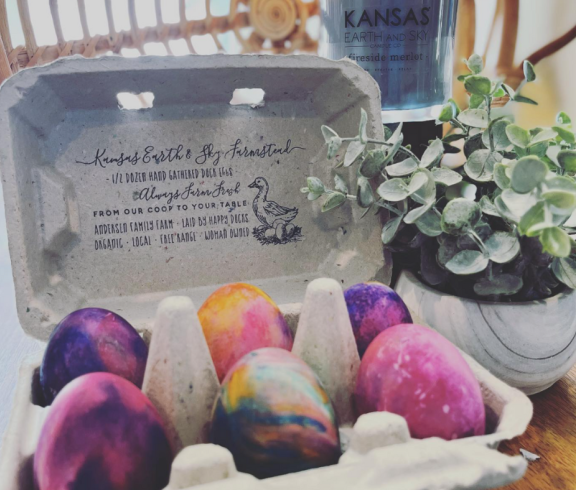 Half a dozen large pekin duck eggs that have been dyed rainbow colors for Easter, in a recycled paper egg carton. A stamp on the carton reads, "Kansas Earth & Sky Farmstead 1/2 dozen hand gathered duck eggs. Always farm fresh. From our Coop to your Table." 